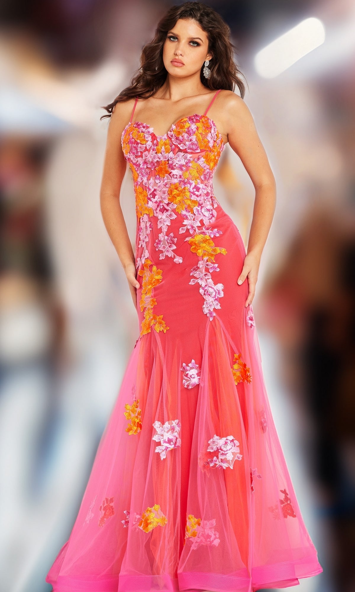 A woman poses on a blurred background wearing the Long Prom Dress 36843 by Jovani, a pink strapless gown with orange and white floral embroidery and a sweetheart neckline.